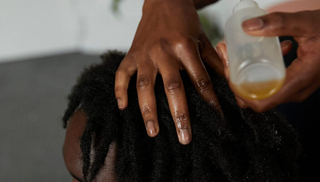 Young black man with locs gets his hair oiled by friend whilst sitting on the bedroom floor