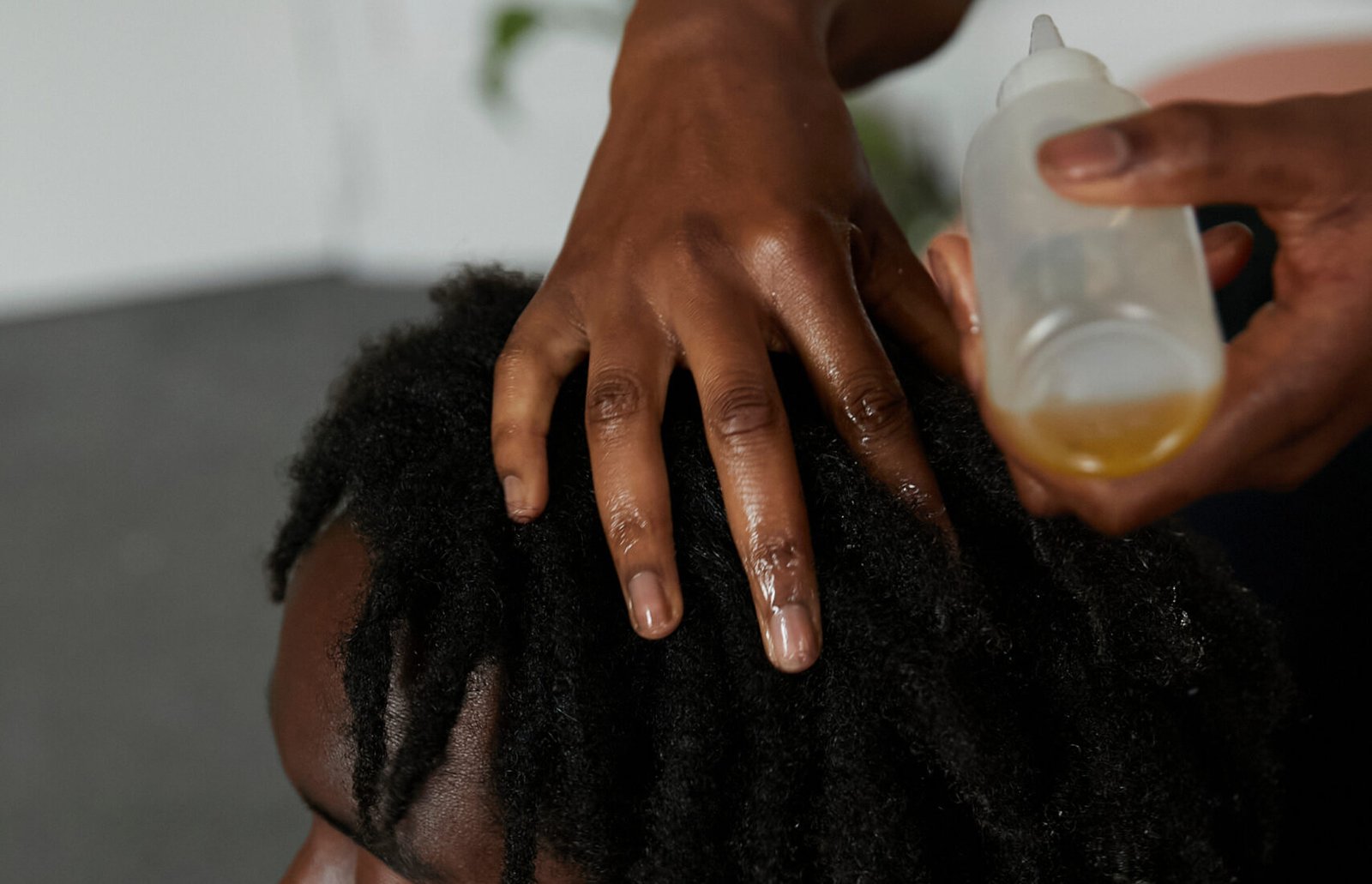 Young black man with locs gets his hair oiled by friend whilst sitting on the bedroom floor