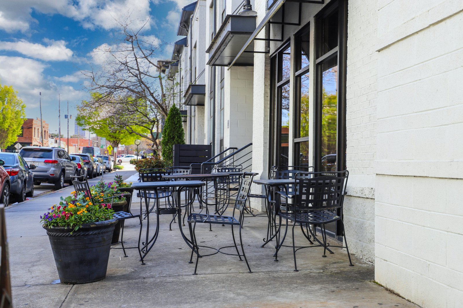 black metal tables and chairs in an outside dinning area at a restaurant in front of a white building with lush green trees and cars parked along the street and black flower pots with colorful flowers black metal tables and chairs in an outside dinning area at a restaurant in front of a white building with lush green trees and cars parked along the street and black flower pots with colorful flowers